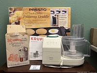 Photo showing Krups food processor, Waring juicer box, and Presto griddle box arranged together against a wall.