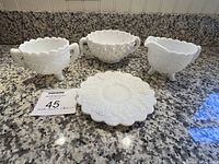 Four white milk glass pieces on granite countertop: three bowls with handles and one round plate with detailed patterns including hobnail and grape cluster designs.
