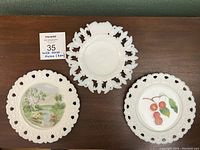 Three vintage milk glass plates displayed on a wooden table including one plain white plate and two hand painted plates, one with cherries and one with a landscape scene, all featuring pierced rims.