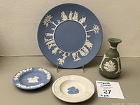 Large blue Wedgwood Jasperware plate with circular white relief decorations, smaller blue plate with white reliefs, white and blue ashtrays, and green Jasperware vase with white relief, displayed against a neutral background.