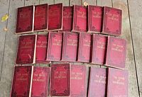 Photo of all 20 volumes of The Book Of Knowledge laid out on a wooden surface showing red hardcovers with black decorative borders and titling, condition worn.