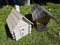 Photo showing both the white log cabin style bird house and larger wooden bird feeder together on grass outdoors.