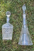Two clear crystal decanters with stoppers shown lying on grass; different sizes and detailed cut glass designs visible