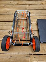 Foldable hand trolley showing metal frame, red wheels, and orange elastic cords on wooden floor