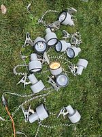 Twelve mostly white clip-on lights with spiral cords arranged on grass, some lit, showing adjustable heads and clamp clips.