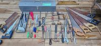 Long metal toolbox and various hand tools laid out on wooden surface.