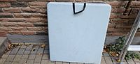 Photo of folded rectangular white plastic folding table with black handle, showing the top and sides against a brick background.