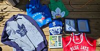 Overview of apparel including two Toronto Blue Jays hoodies, Toronto Maple Leafs apparel and Green Bay Packers framed memorabilia along with clocks.