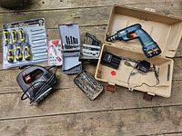 Photo showing cordless drill in case with charger, multiple drill bit sets in plastic and metal containers, Skill jigsaw with cord, and new packs of screwdrivers.