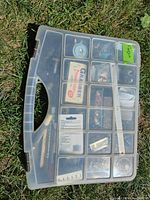Top view of transparent tool case with multiple compartments containing various small hardware and electronic parts, including a grabber tool package visible.