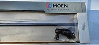 Close-up of Moen towel bar in original packaging showing oil rubbed bronze finish, part of the packaging box with Moen branding and Bradshaw series name visible.