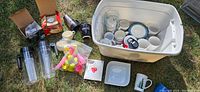 Tub filled with various kitchenware items including cups, mugs, plates, jugs, and other kitchen utensils overseen on grass.