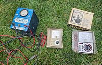 Photo showing all three items together on grass: blue Motomaster charger with attached cables, beige Heathkit multitester, and Micronta multimeter in bubble wrap and original box.