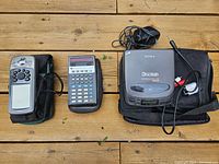 Three vintage electronic devices arranged on wooden surface: Garmin GPS with worn case, TI SR-50 calculator with black case, Sony Discman portable CD player with case and cables