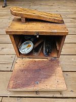 Wooden box with door open showing contents inside: round Dubbin tin, brushes, and tube, with wooden shoe-shaped footrest on top.