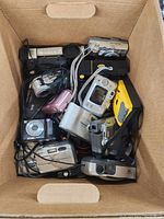 One top-down image showing a cardboard box filled with various small cameras, camera accessories, and charging cables.