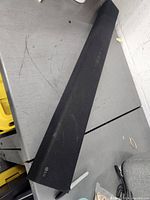 Front view of the LG sound bar placed diagonally on a gray surface, showing overall length and black fabric covering with slight wear.