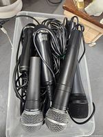 Photo shows a plastic container with seven wired microphones in a jumble including brands Shure and Guitar Hero, with cables tangled.