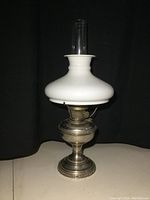 Full view of vintage Aladdin metal oil lamp with white milk glass shade and glass chimney on a light-colored surface with black background.
