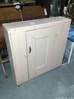 Front view of the rectangular pale painted wood wall cabinet with one door, showing aged chipped paint and wear.