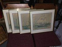 Four framed prints by Ben Babelowsky, each featuring tall ships, arranged on a chair; frames are large with thin gold edges; one frame missing glass