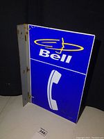 Photo showing one side of the Bell Telephone double-sided reflective flange metal sign standing on a white surface with black backdrop.