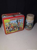 Front view of embossed Disney Express metal lunchpail with colorful Disney characters and matching thermos next to it