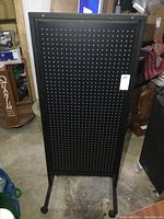 Front view of the black metal peg board display on wheels, showing the evenly spaced holes in the peg board and the metal frame base with caster wheels.