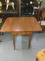 Front view of antique drop leaf table with drawer closed and leaves down in a room with assorted furniture.
