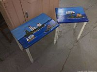 Both hand painted wood tables showing blue nautical scenes with ships and houses, cream turned legs, on cement floor.