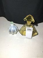 Photo showing two glass perfume bottles on a white surface with black background: a smaller white-blue frosted bulbous bottle and a larger amber-yellow geometric bottle with stopper.