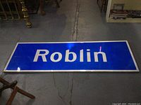 Front view of large rectangular highway sign with white reflective letters Roblin on blue background.