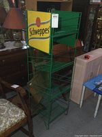 Green metal Schweppes branded wire store rack with yellow signage and multiple shelves shown from front angle.