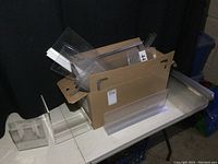 Box filled with various sizes of clear plastic sign holders stacked inside and next to box on table surface.