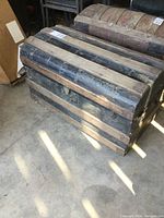 Front and top angled view of an antique trunk showing black wood with metal bands reinforced on exterior. Hinges disconnected from lid, handles missing. Aged patina and wear.