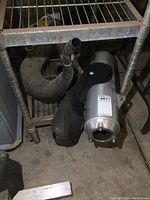 Three exhaust components on metal rack and floor: Kawasaki Ninja stock muffler and two ATV exhaust pipes, one curved black and another smaller black.