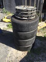Stack of four winter tires mounted on steel wheels with hubcaps displayed outdoors.