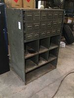 Front view of metal parts cabinet showing 18 drawers and 6 open cubby compartments with surface rust on the bottom and edges.