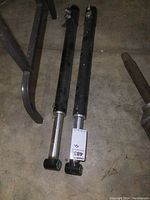Pair of black hydraulic shocks lying on concrete floor showing length and end eyelet attachments.