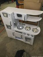 Front and right side view of the white childs kitchen play center with various features including fridge, microwave, stove, sink, and shelving.