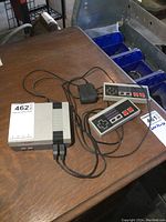 Nintendo NES console with two wired controllers and power adapter on a wooden table, showing front and side views of the unit.