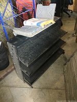 Black metal parts shelf angled with three tiers, showing wear and a parts book in metal holder on top.