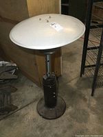 Full view of the patio heater showing the round metal base, cylindrical propane canister housing, and large heat reflector on top.