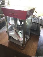 Front and side view of the popcorn machine showing red top and glass sides on wooden table in an indoor space, with black power cord visible.