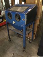 Front angled view of the blue sandblasting cabinet with viewing window and glove ports visible, mounted on a metal frame with wheels.