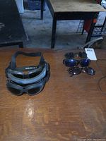 Photo showing three pairs of goggles with black straps stacked, and three pairs of sunglasses with different colored lenses arranged on table