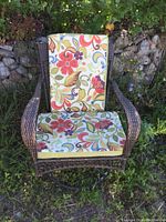 Front view of the dark brown plastic wicker swivel chair with floral cushions placed outdoors against rocks and greenery.