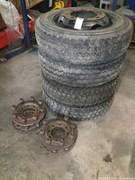 Four stacked truck tires mounted on 19.5 inch steel wheels with tread visible. Four rusty metal wheel conversion spacers with lug nuts laid on floor nearby.