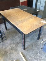View of the wooden table from an angle showing the plywood top and black painted legs with wear and paint chipping.