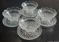 Four clear pressed glass dessert bowls with matching saucers arranged on a surface showing diamond-patterned texture and scalloped edges of bowls.